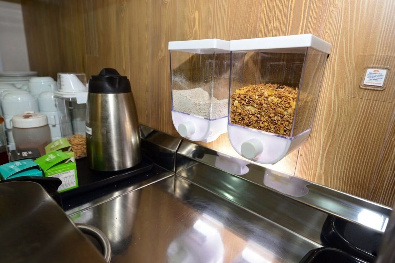 Een keukentafel met een roestvrijstalen koffiepot, twee doorzichtige dispensers met poeder en granola, en een houten wand.