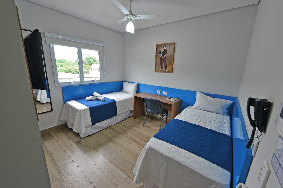 Ein Zimmer mit zwei Betten, einem Schreibtisch, einem Stuhl, einem Deckenventilator und einem Fenster.