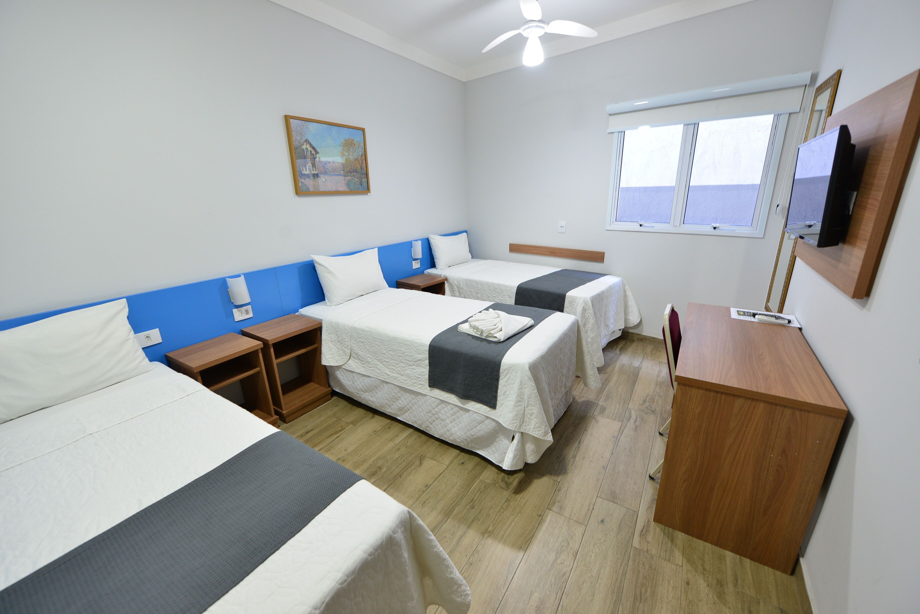 A hotel room with two beds, a desk, a TV, a window, and a painting on the wall.
