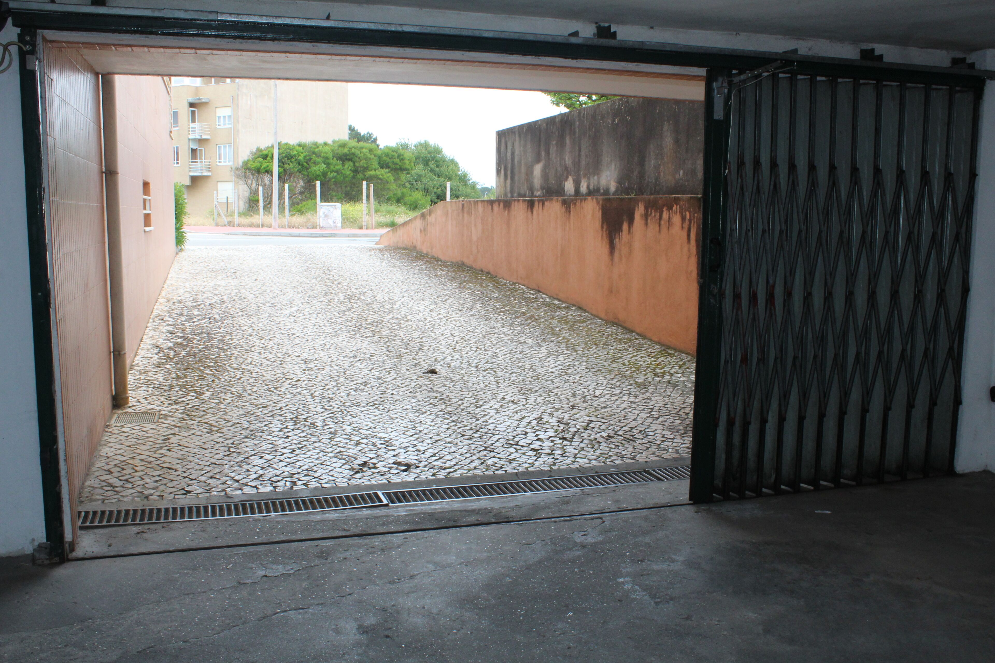 On aperçoit, à travers une grille métallique, une zone pavée avec un mur en béton et un bâtiment en arrière-plan.