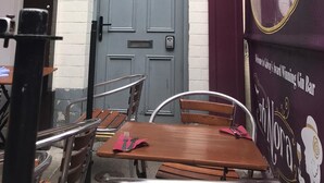 Uma área externa com mesas e cadeiras, uma porta cinza fechada e uma parede com um mural anunciando um bar de gin.
