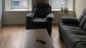 Sala de estar com sofá, poltrona, mesa de centro e controle remoto. Duas janelas oferecem vista para o exterior, uma estante com objetos decorativos e um abajur pendurado no teto.
