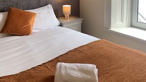 Cama bem arrumada com manta marrom e toalha branca, luminária de cabeceira e janela com persianas brancas.