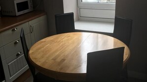 Uma pequena área de refeições com uma mesa redonda de madeira e quatro cadeiras, um micro-ondas na bancada e uma janela com vista para edifícios.