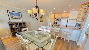 A modern kitchen with white cabinets, a central island, and a dining area with a glass table and white chairs. - Queen's Grant E-117 – Perfect Beach Getaway (Topsail Beach)