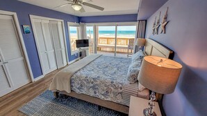 A bedroom with a bed, a TV, a ceiling fan, and a view of the beach. - Queen's Grant E-117 – Perfect Beach Getaway (Topsail Beach)