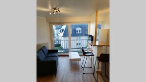 A modern apartment with a wooden floor, a black sofa, a dining table with two chairs, and a large window offering a view of neighboring buildings. - Appartement Le Conquérant - Caen (Caen)