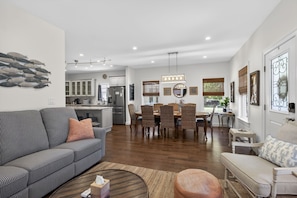 A modern living room with a sofa, ottoman, dining table with chairs, kitchen area, and a staircase.