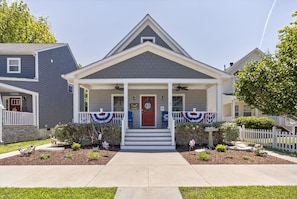 A two-story house with a front porch, a red door, and a small front yard with a bird bath and decorative flags.