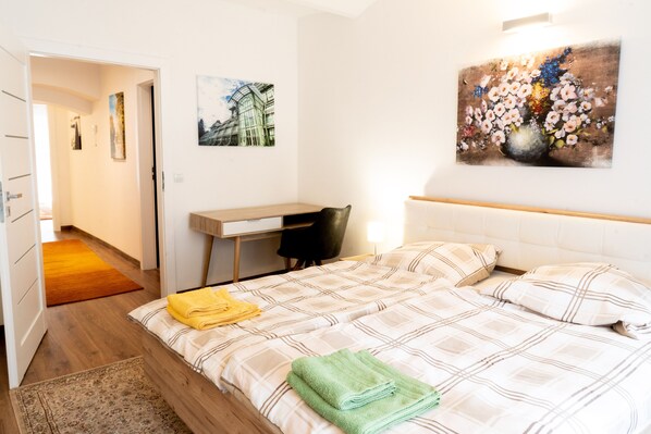 A hotel room with a bed, a desk, and a chair. There is a painting on the wall and a door leading to another room.