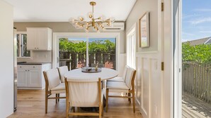 Une cuisine moderne avec des meubles blancs, une table de salle à manger ronde entourée de chaises, et une grande fenêtre donnant sur une terrasse en bois et un espace verdoyant.