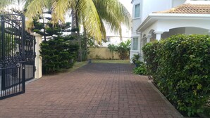 A paved driveway leading to a gated entrance with a white building on the right and a palm tree on the left. - Villa The Golden Dream  (Flic en Flac)