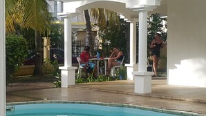 A pool area with a covered seating area, palm trees, and a building in the background. - Villa The Golden Dream  (Flic en Flac)