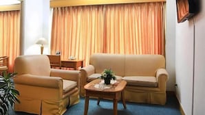 A hotel room with a beige sofa, a matching armchair, a small wooden table with a plant, a desk with a lamp, and a TV mounted on the wall. - Hotel Marcopolo (Bandar Lampung)