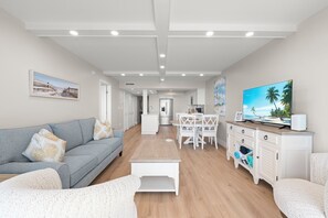 Una sala de estar moderna con un sofá gris, sillones blancos, una mesa de centro de madera y un televisor sobre un mueble blanco.