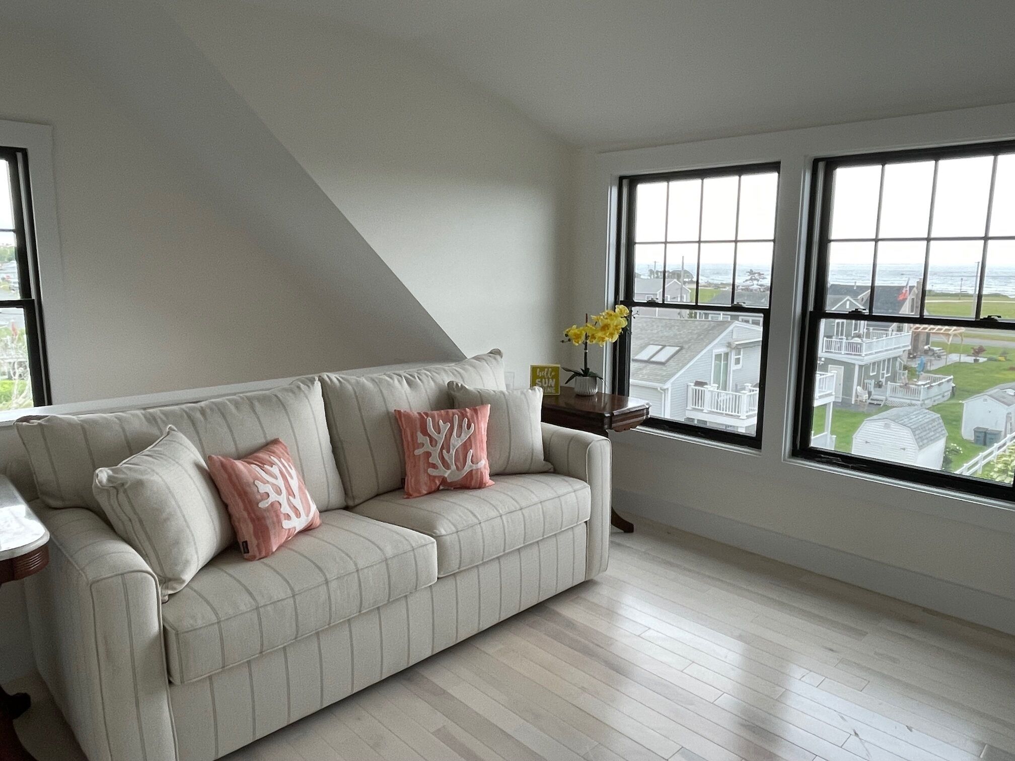 A modern living room with a light-colored sofa, two windows with a view of houses, and a small table with a vase of flowers.