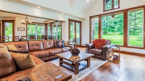 A spacious living room with a large sectional sofa, a coffee table, and a fireplace. There are large windows offering a view of the outdoors.