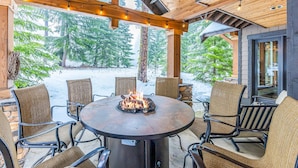 A covered outdoor seating area with a fire pit, wicker chairs, and a view of a snowy forest.