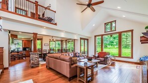A spacious living area with a high ceiling, wooden flooring, a large sofa, and a dining area visible through an open doorway.