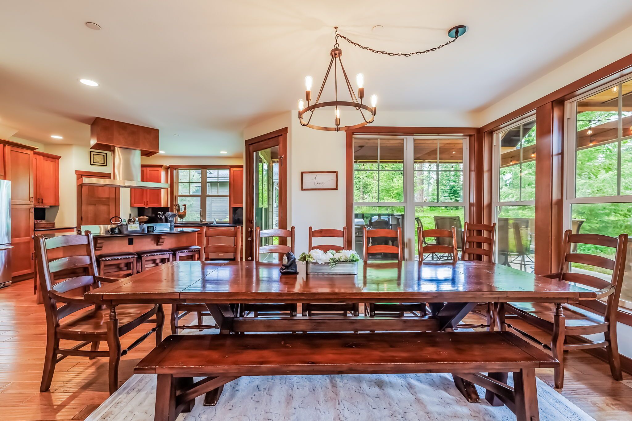 A spacious dining area with a large wooden table and chairs, a chandelier, and large windows offering a view of the outdoors.