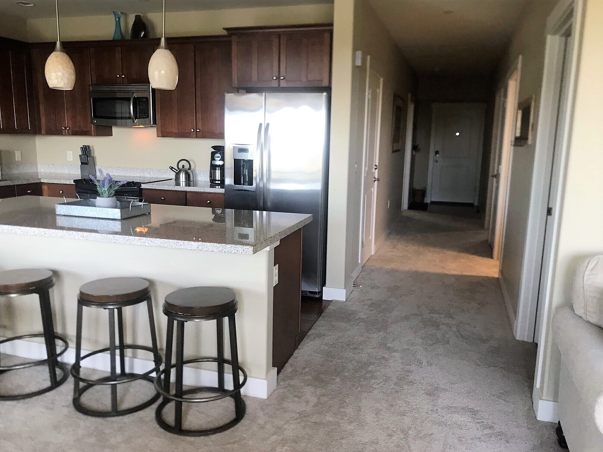 A kitchen with dark wooden cabinets, a stainless steel refrigerator, a microwave, and a coffee maker. There are three stools at the kitchen island.
