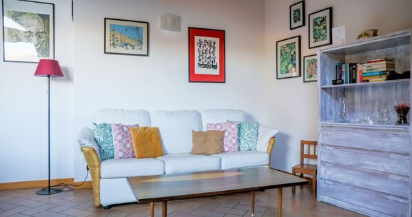 Una sala de estar con un sofá blanco, cojines coloridos, una lámpara roja de pie y una estantería con libros y objetos decorativos.