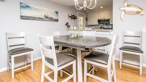 A dining area with a round table surrounded by white chairs with grey cushions, a chandelier above, and a painting of a boat on the wall.
