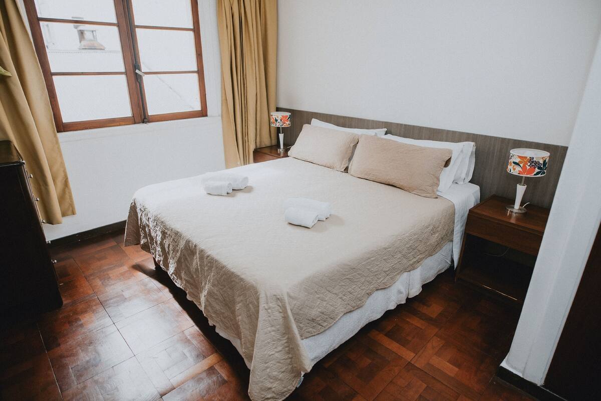 Un lit bien fait, avec une couette blanche et deux oreillers, une table de chevet en bois avec une lampe, et une fenêtre avec des rideaux.
