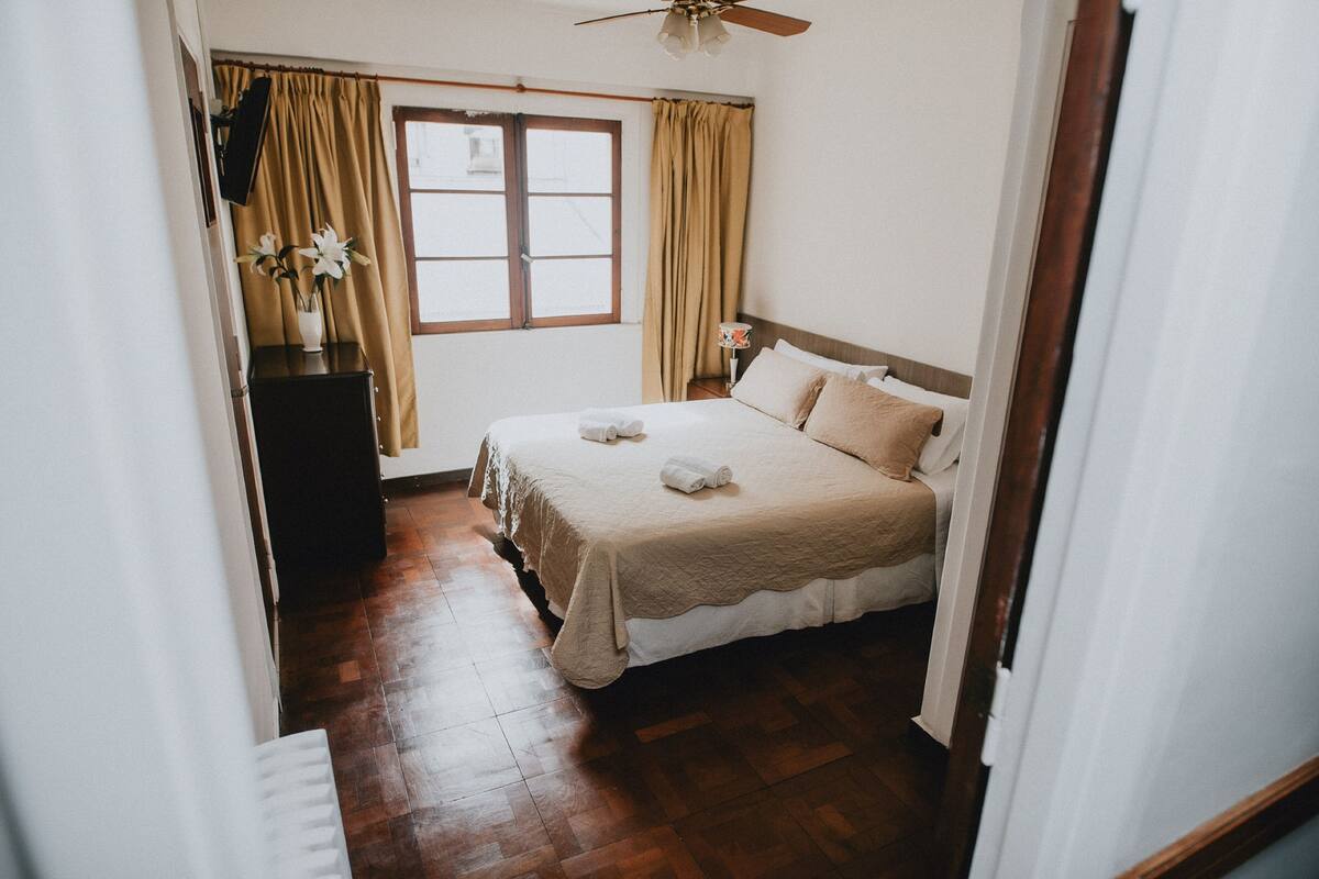 Une chambre à coucher avec un lit, une table de chevet, une fenêtre avec des rideaux et un ventilateur de plafond.