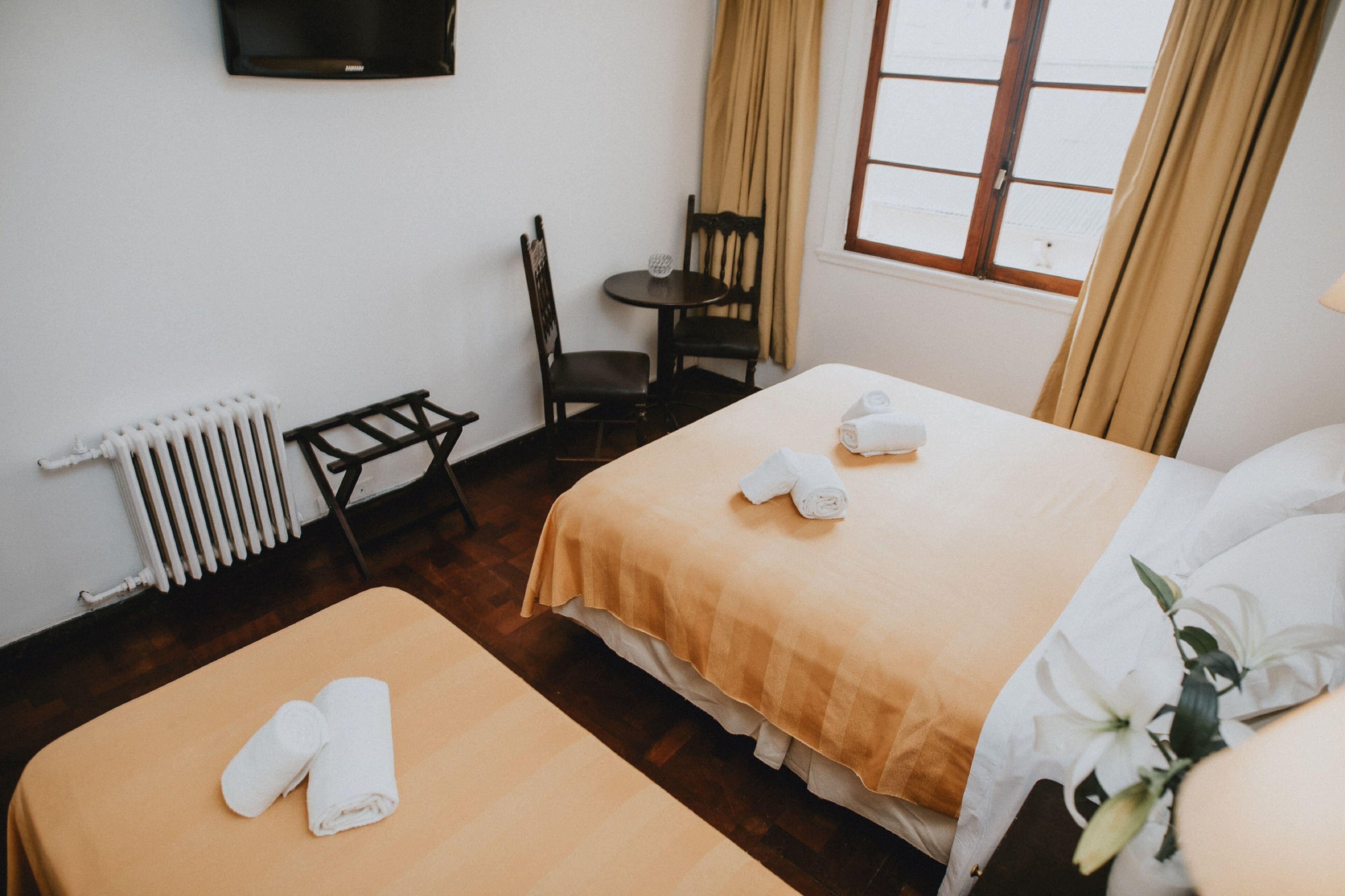 Habitación de hotel con dos camas, una mesita, una silla, televisor montado en la pared y una ventana con cortinas.