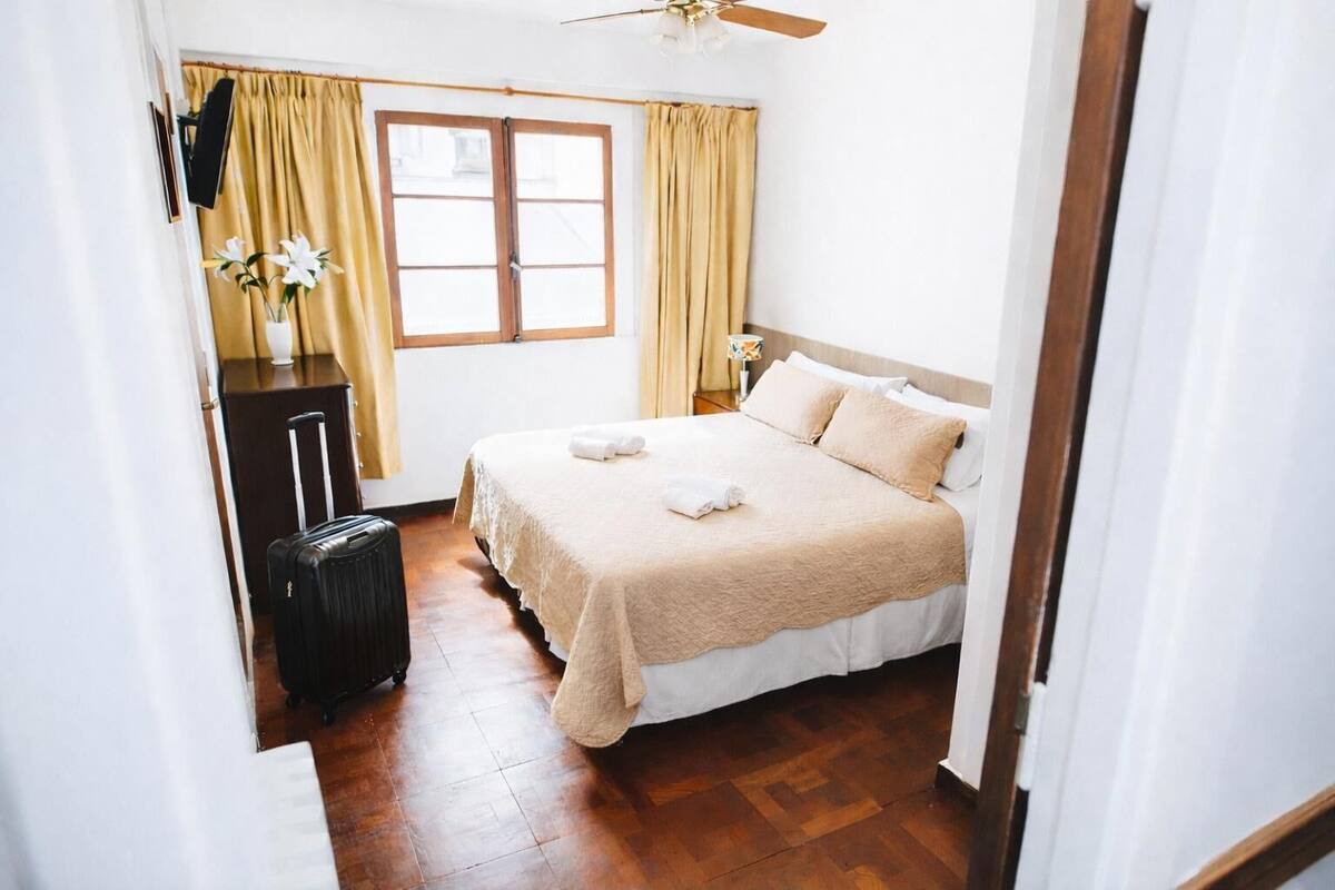 Une chambre d’hôtel avec un lit, une valise, un bureau et une fenêtre avec des rideaux.