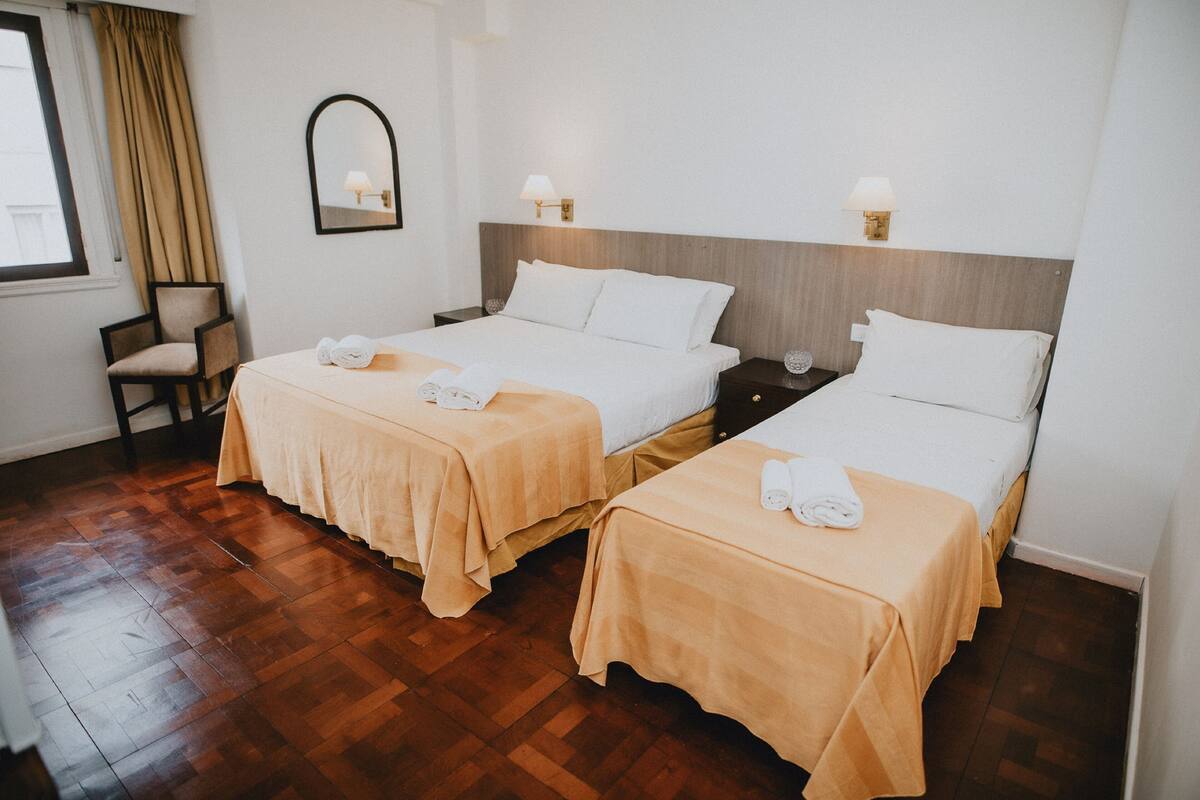 Une chambre d’hôtel avec deux lits, un plancher en bois, une chaise et une fenêtre avec des rideaux.