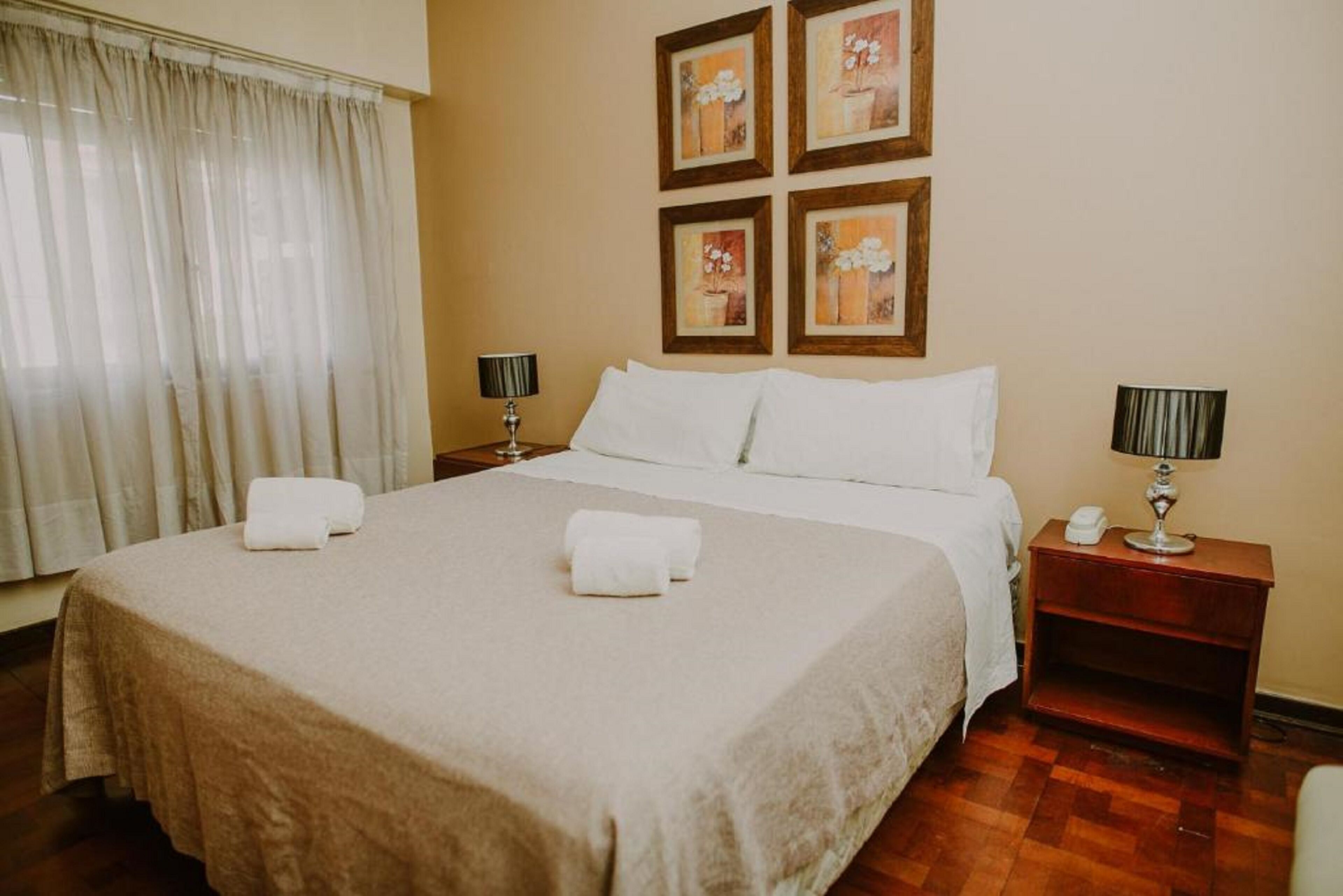 A neatly made bed with white linens and two pillows, a bedside table with a lamp, and a wall with four framed floral paintings.