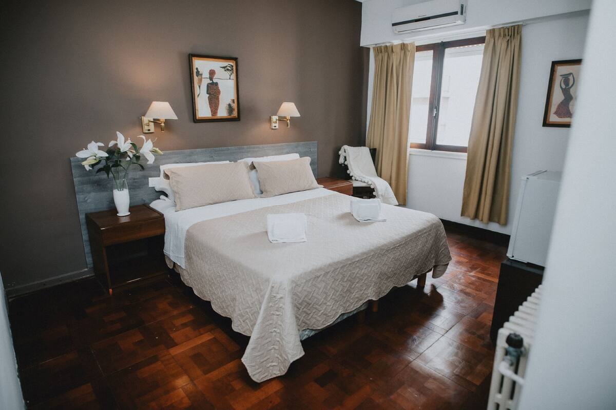 Un lit bien fait, recouvert d’une courtepointe blanche et agrémenté d’oreillers, une table de chevet avec un vase de fleurs et une lampe fixée au mur.