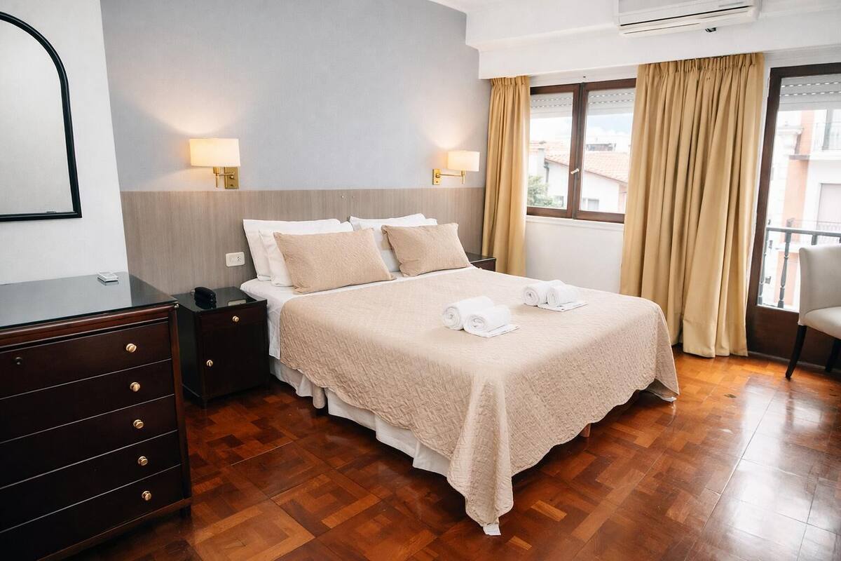 Une chambre d’hôtel comprenant un lit, des tables de chevet, une commode, une fenêtre avec des rideaux et une vue sur les bâtiments environnants.