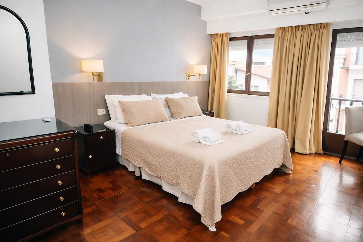 A hotel room with a bed, bedside tables, a dresser, a window with curtains, and a view of buildings outside.