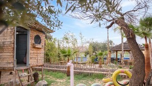 Une structure rustique en bois, dotée d’une porte, d’une échelle et d’une zone délimitée par une clôture, où se trouvent des pneus et des palmiers.