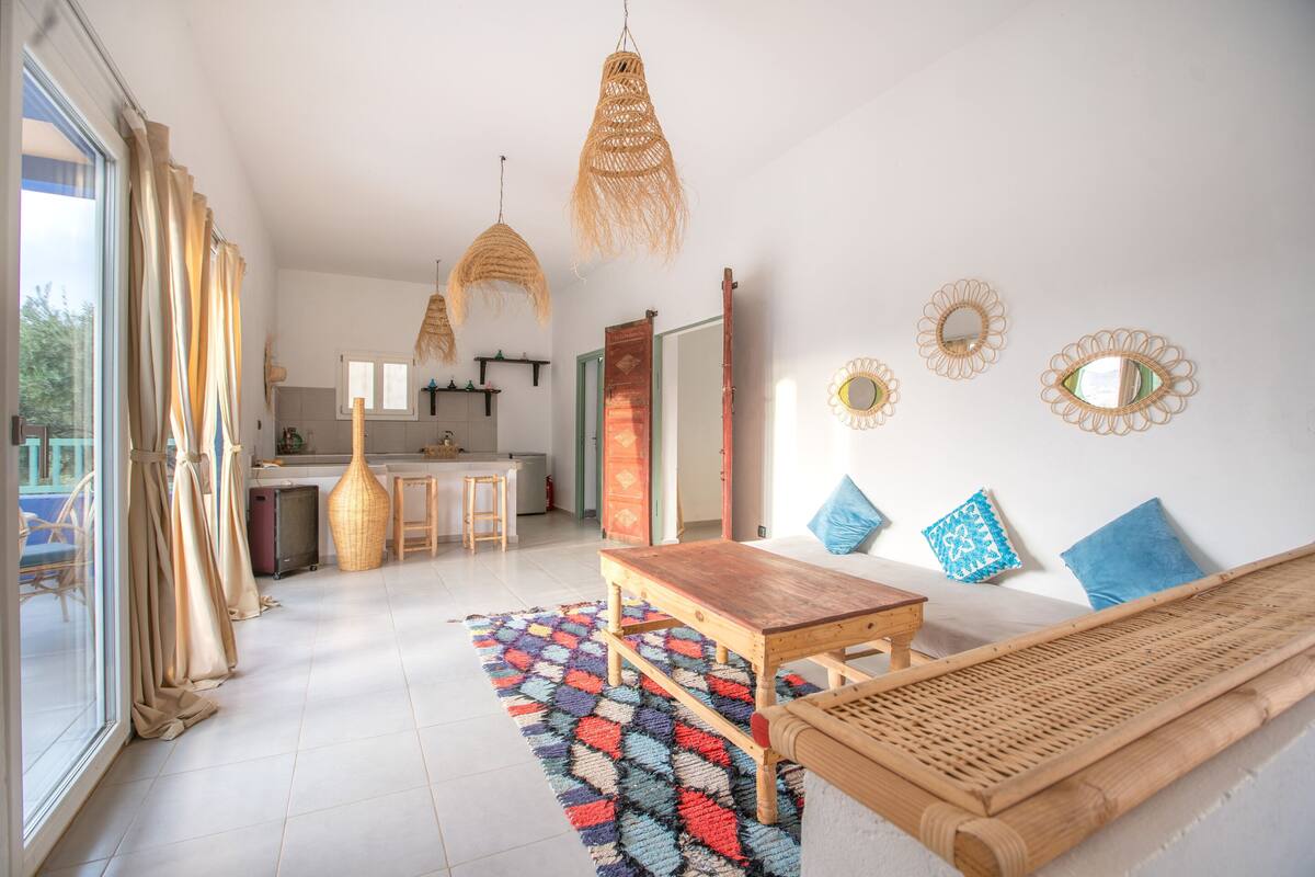 Un vaste espace de vie avec une table et un banc en bois, un tapis coloré et une cuisine en arrière-plan.
