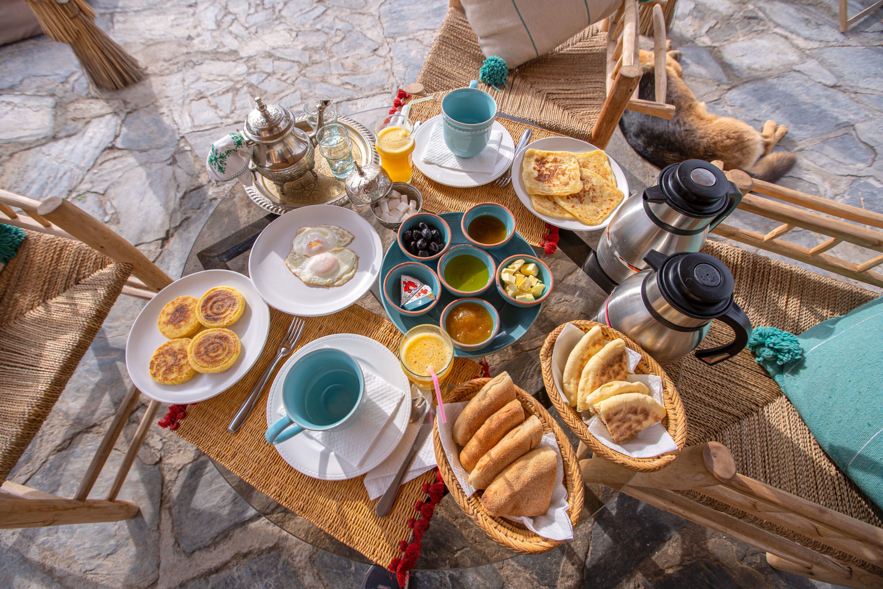 A breakfast setup with plates of food, cups, and a teapot on a woven mat.