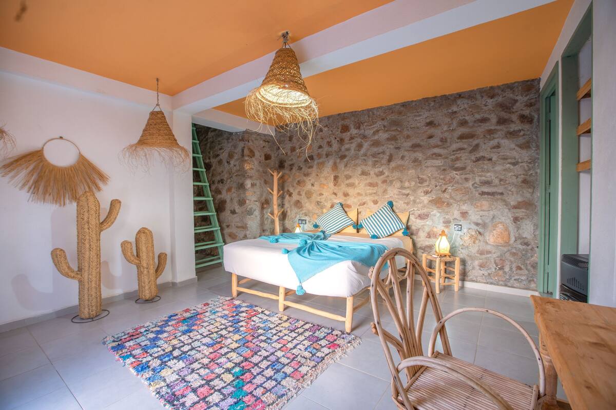 Une chambre avec un mur en pierre, un lit avec une literie bleue, un tapis coloré et un luminaire suspendu.