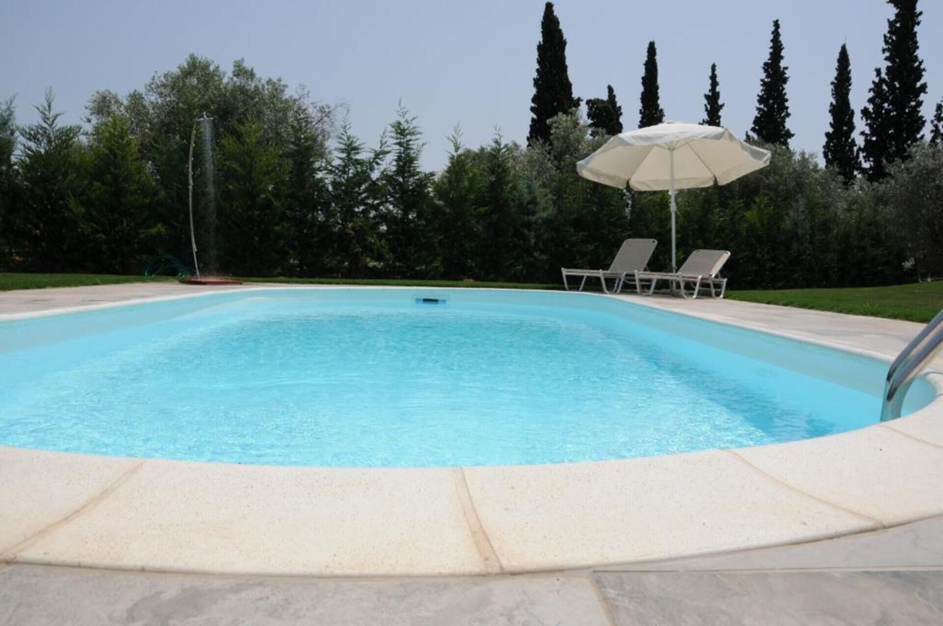 Une piscine rectangulaire entourée d’une terrasse de couleur claire. On y trouve deux chaises longues et un parasol près de la piscine.