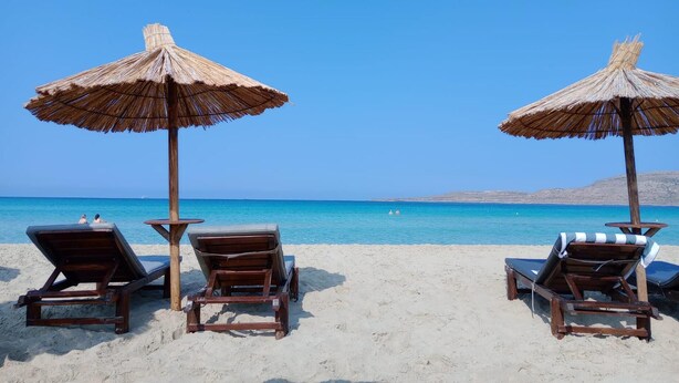 Dos sillones de madera con sombrillas de paja en una playa arenosa, mirando un mar tranquilo bajo un cielo despejado.