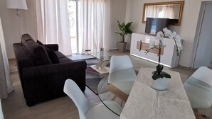Un moderno salón con un sofá marrón oscuro, una mesa de centro de cristal y sillas blancas. Hay un armario blanco con un televisor y una planta encima. Se ve una escalera al fondo.