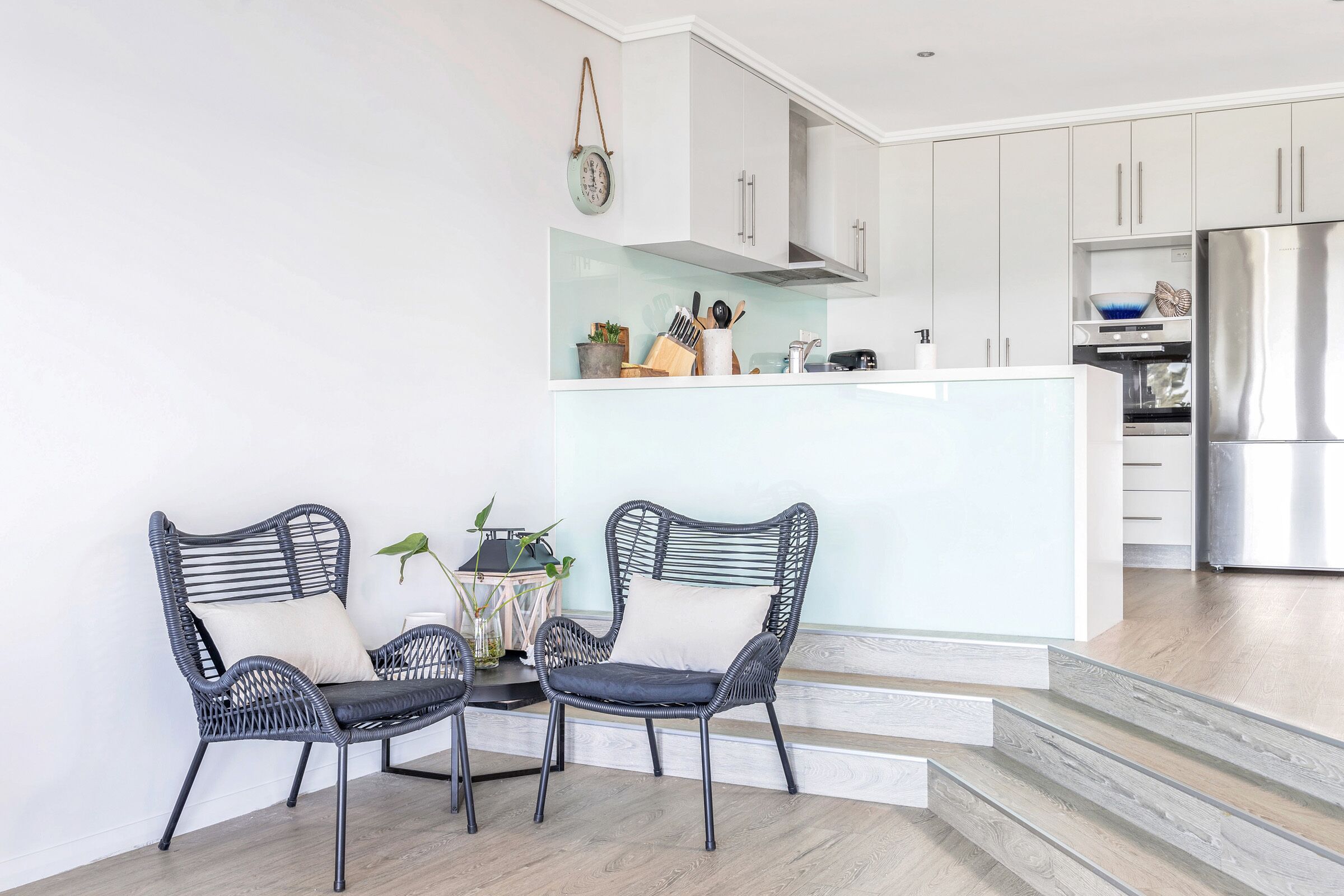 A modern kitchen with white cabinets, a stainless steel refrigerator, and a dining area with wicker chairs.