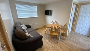 Ein gemütlicher Wohnraum mit einer dunkelgrauen Couch, einem Holzesstisch mit vier Stühlen, einem an der Wand befestigten Fernseher und einem Fenster mit Jalousien.