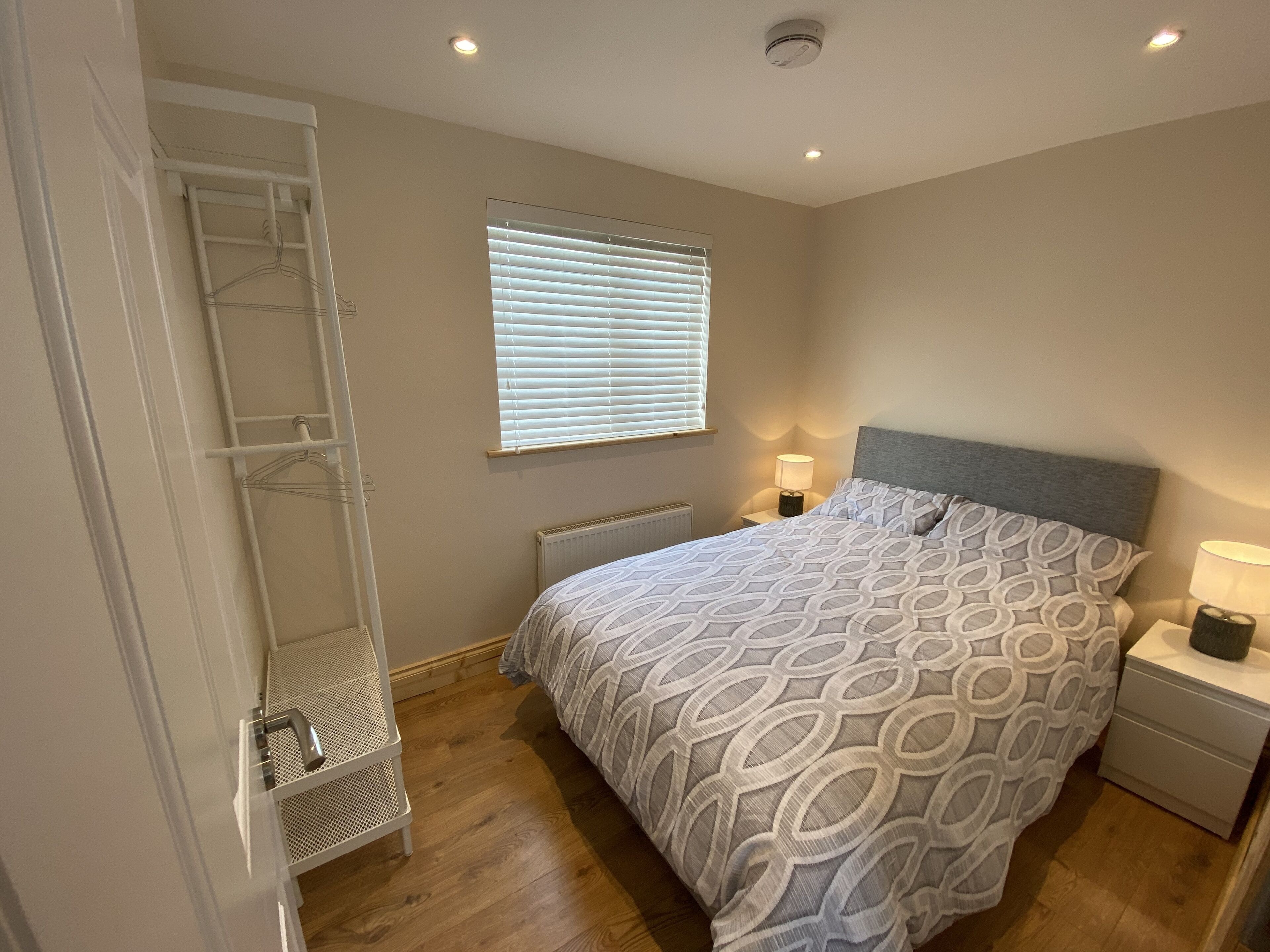 A bedroom with a bed, bedside tables, a radiator, a window with blinds, a hanging wardrobe, and a ceiling light.