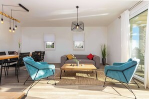Un salon moderne avec une table basse en bois, deux fauteuils bleus, un canapé gris et un coin repas comprenant une table en bois et des chaises noires.