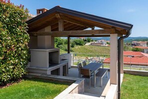 Un patio al aire libre con una parrilla, una zona de estar y vistas a viñedos y edificios al fondo.