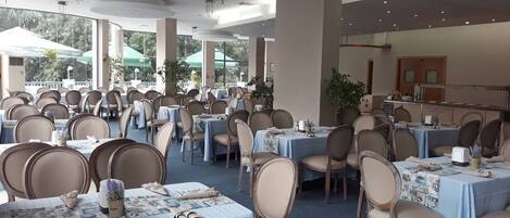 Une salle de banquet avec des tables rondes et des chaises, de grandes fenêtres et un plafond orné de luminaires suspendus.