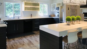 A modern kitchen with a large island, black cabinets, and a white countertop. There are three potted plants on the island, a built-in microwave, and a coffee machine.